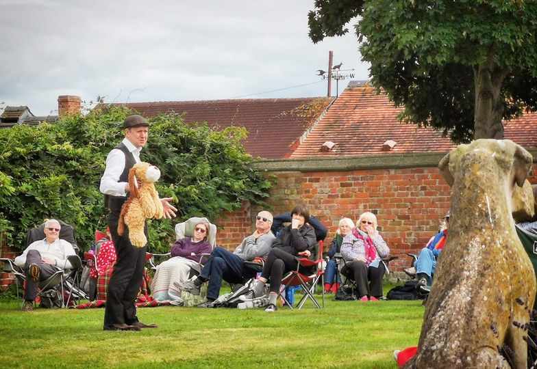 Actor (male) stands in a garden with a toy dog; the audience look on.