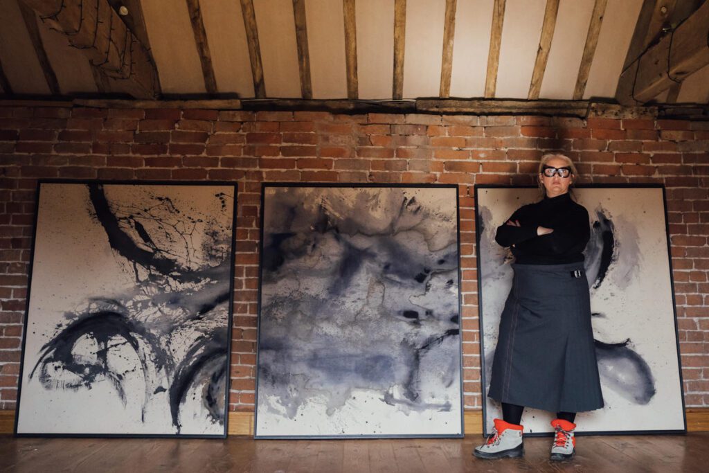 In a brick and timber interior we can see a set of three large canvases. In front of them, the artist who ipresnts as a powerful and defiant woman gressed in black