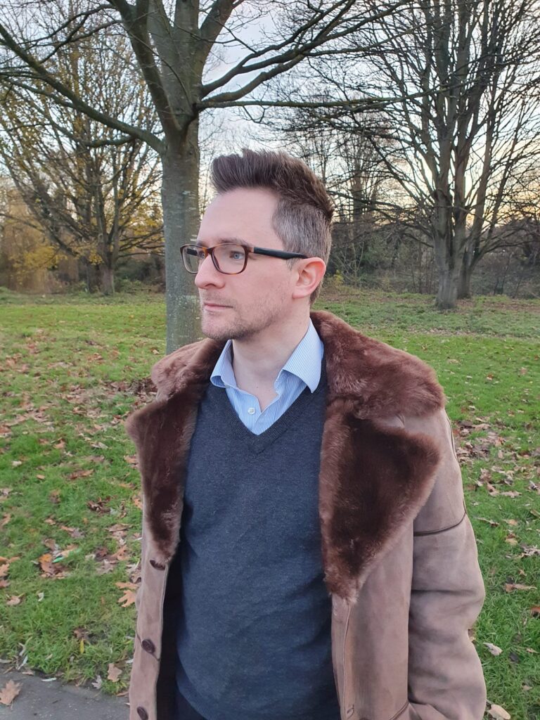 A picture of James Wenn. We can see a male in a blue shirt and a grey v-neck jumper under a brown jacket with a brown fur collar. James is stood in a park in winter and there are trees that are yet to have leaves on them beyond; the sun is low
