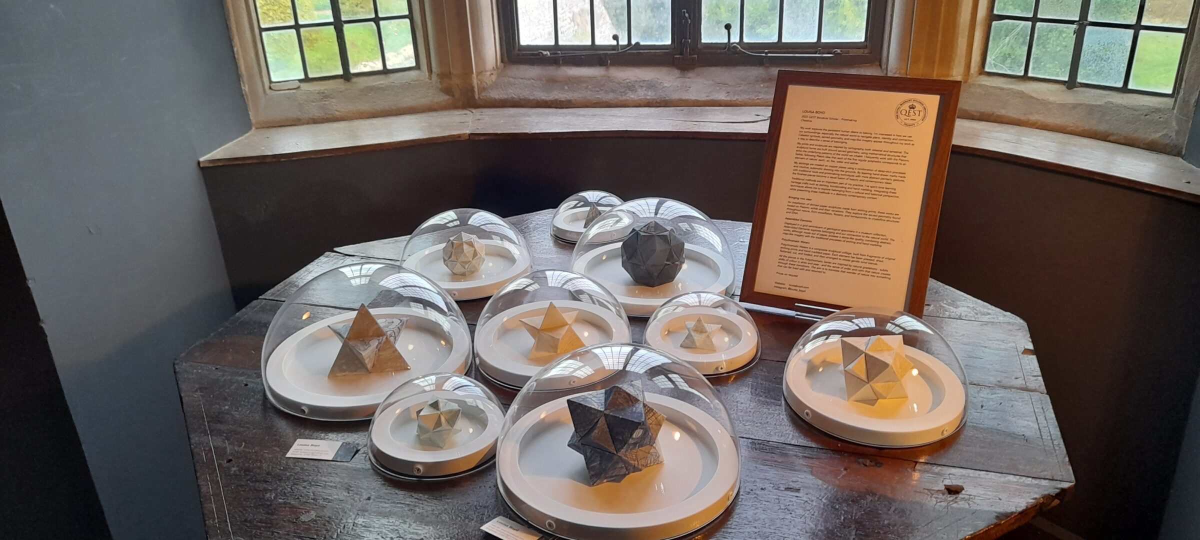 The display features paper sculptures by Louisa Boyd involving stellation, wherein the faces or edges of regular polyhedra are extended outward to form star-like geometric shapes.